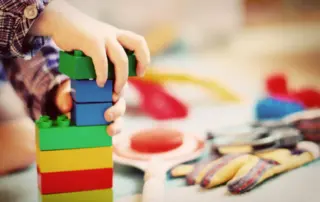 Kleine Hände stapeln bunte Bausteine in einer Kindertagesstätte, während im Hintergrund Spielmaterial liegt.