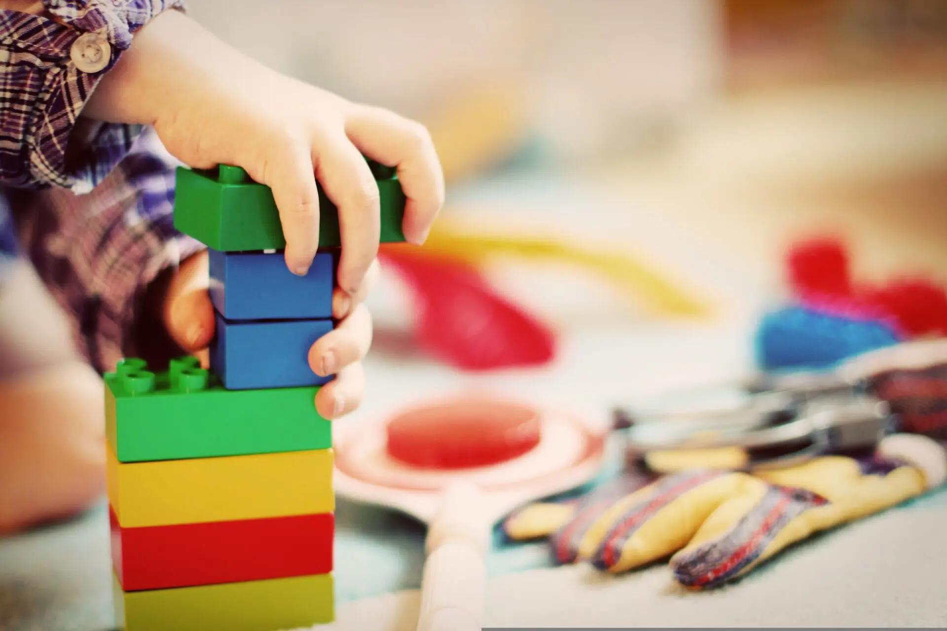 Kleine Hände stapeln bunte Bausteine in einer Kindertagesstätte, während im Hintergrund Spielmaterial liegt.